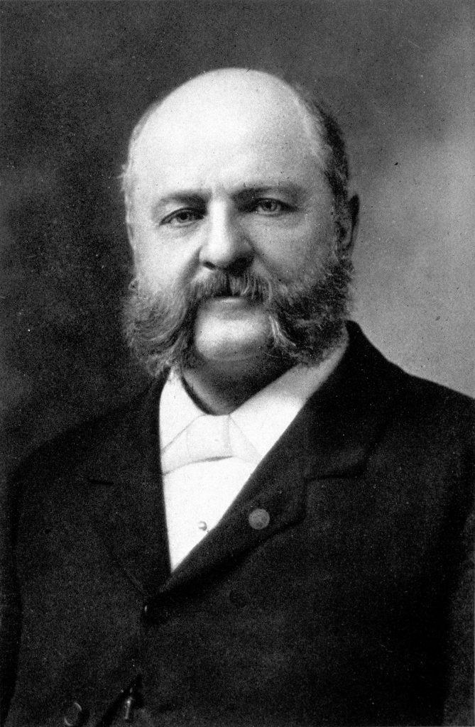 Black and white photo of a bald white man with mutton chop beard
