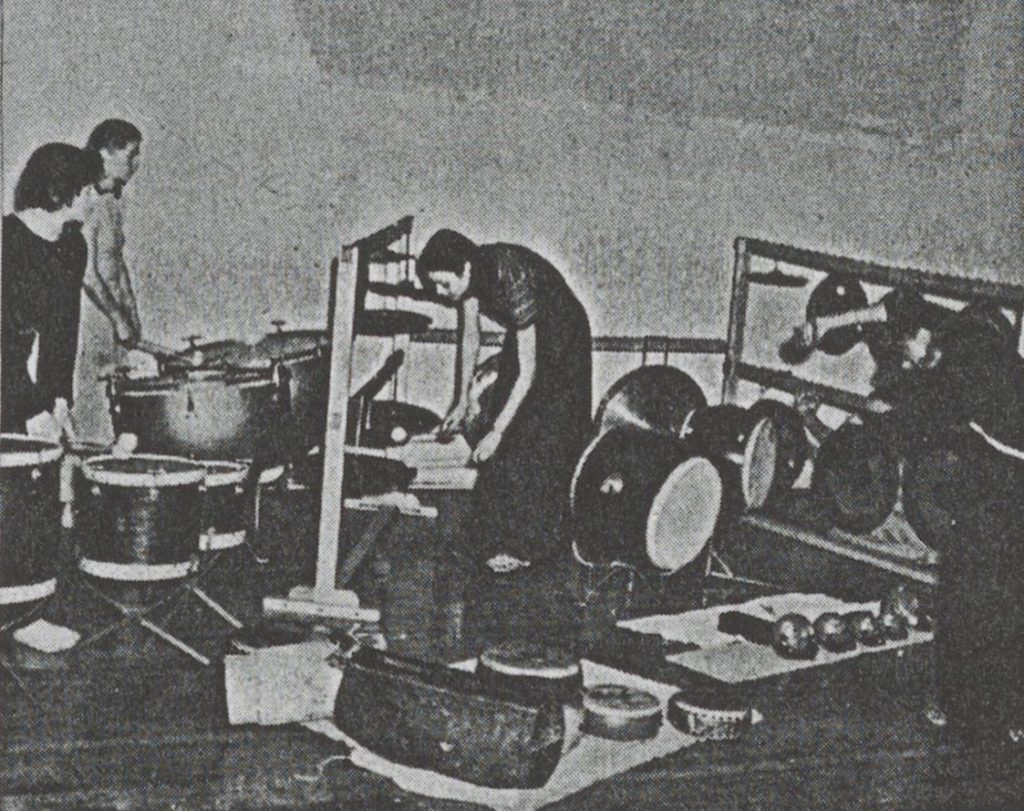 old newspaper photograph of three people in a room with various kinds of drums and percussion instruments