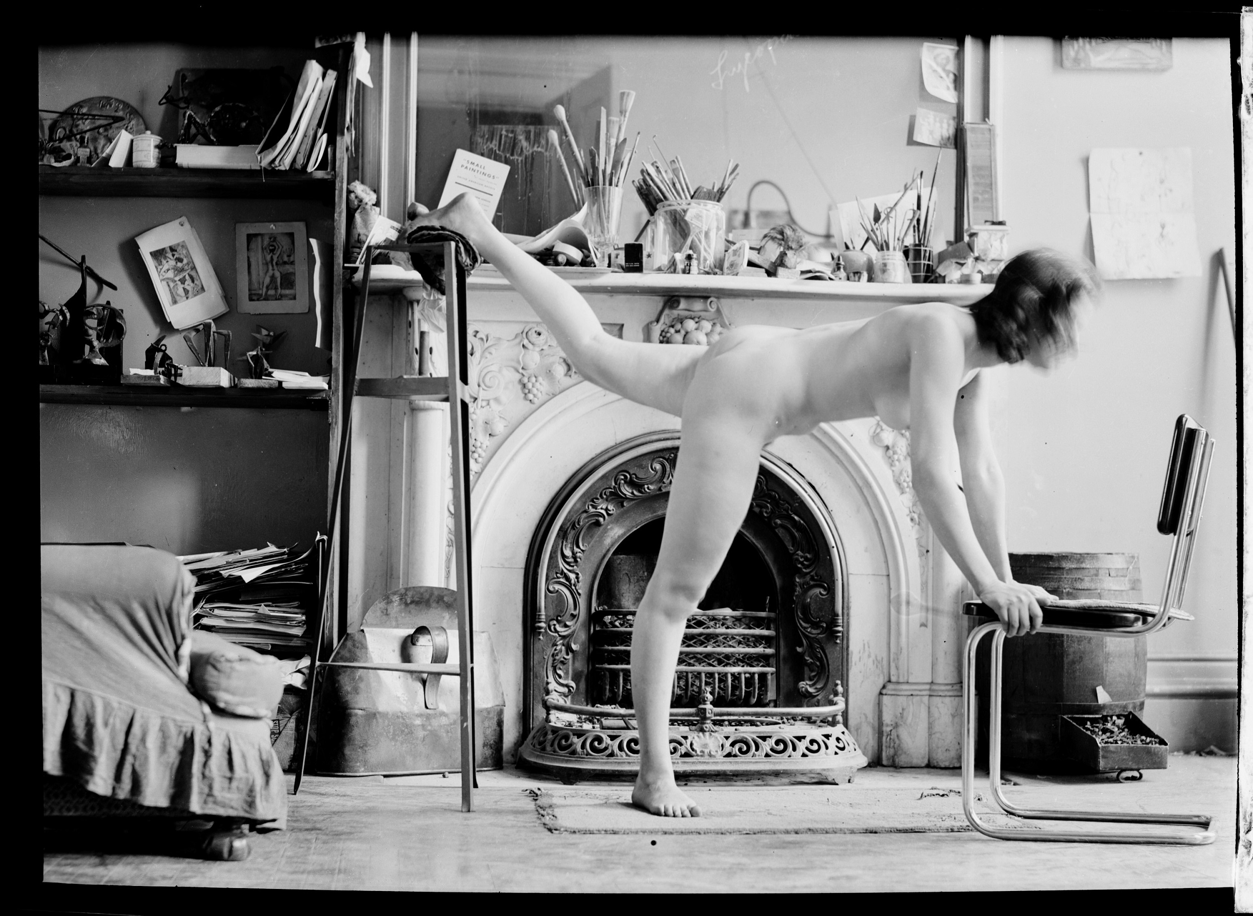 Black and white photograph of a nude woman with her right leg stretched behind her, standing in front of a fireplace