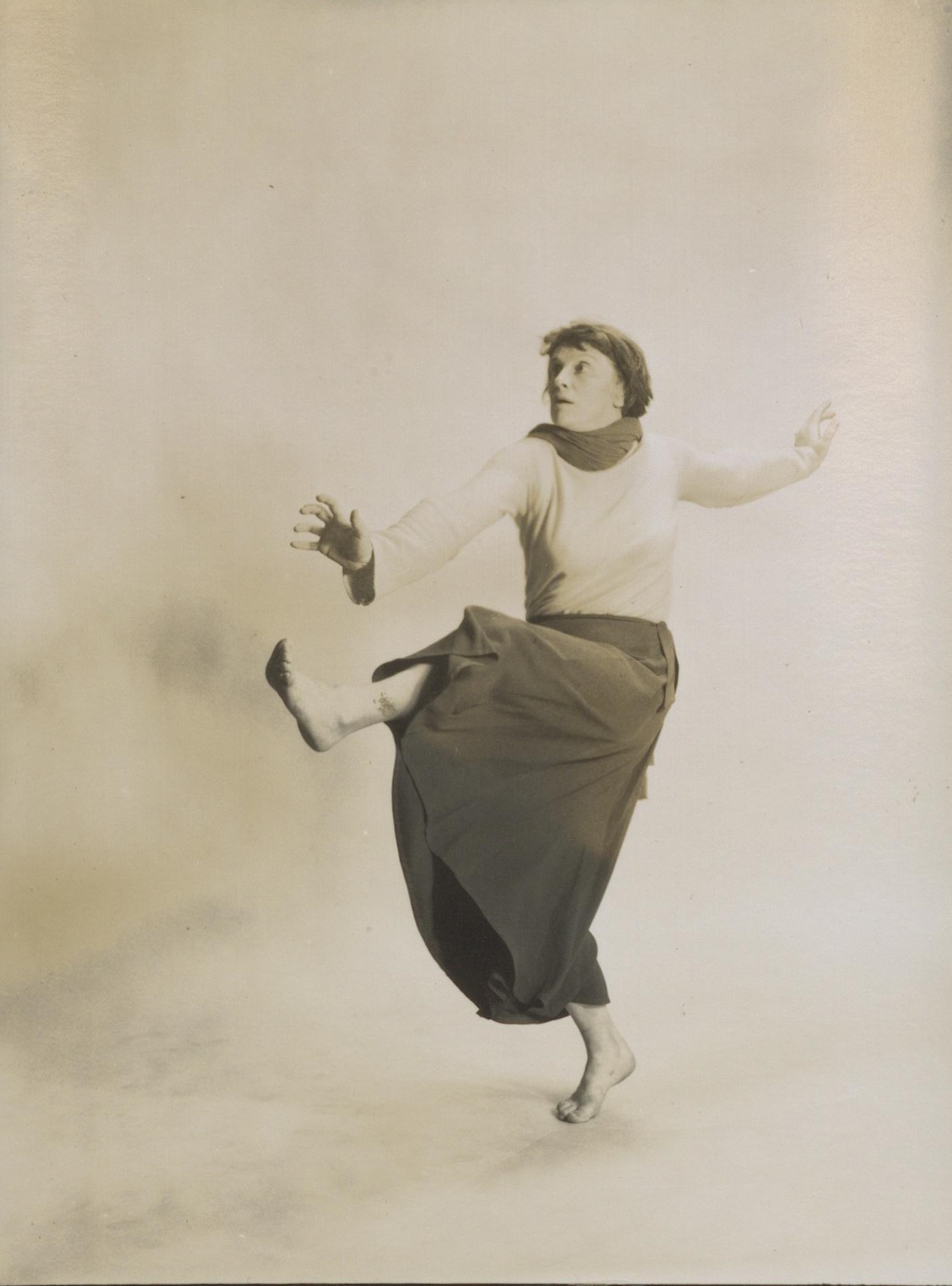 Black and white photograph of a female dancer in a long skirt, kicking her left leg