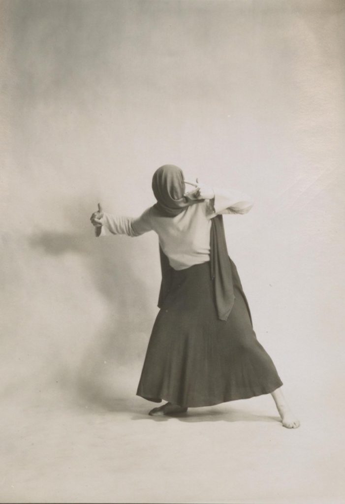 Black and white photograph of a female dancer in a long skirt, a scarf wrapped over her face