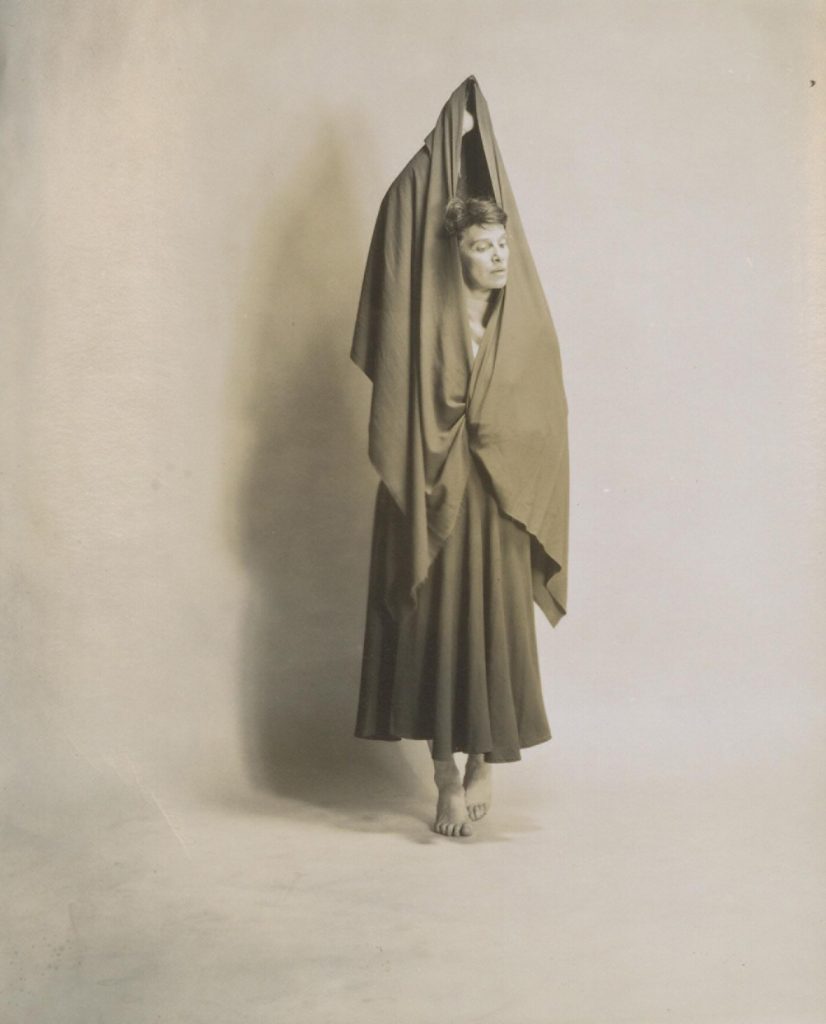 Black and white photograph of a female dancer in a long skirt, hands above her head and holding scarf that drapes over her body