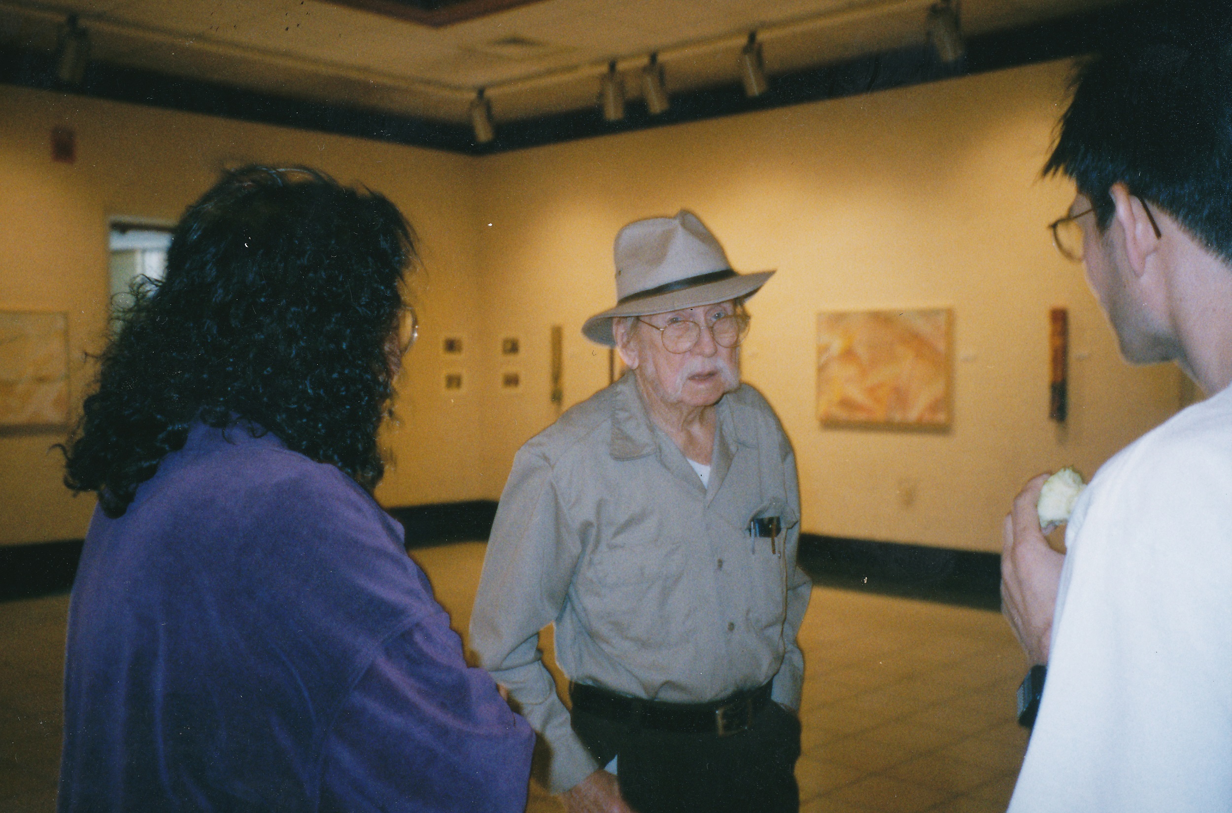 Color photograph of a woman with curly dark hair, wearing a purple velvet top, speaking with an older man with a white moustache, wearing a hat, and a younger man with dark hair and glasses.