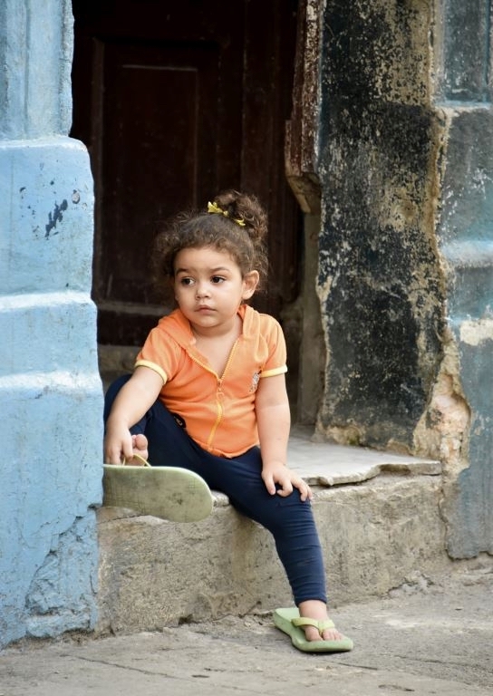 Color photograph of a young girl in an orange shirt and green flip flops sitting in a concrete doorway