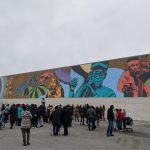 Photograph of square building with colorful mural and a crowd of people looking at it