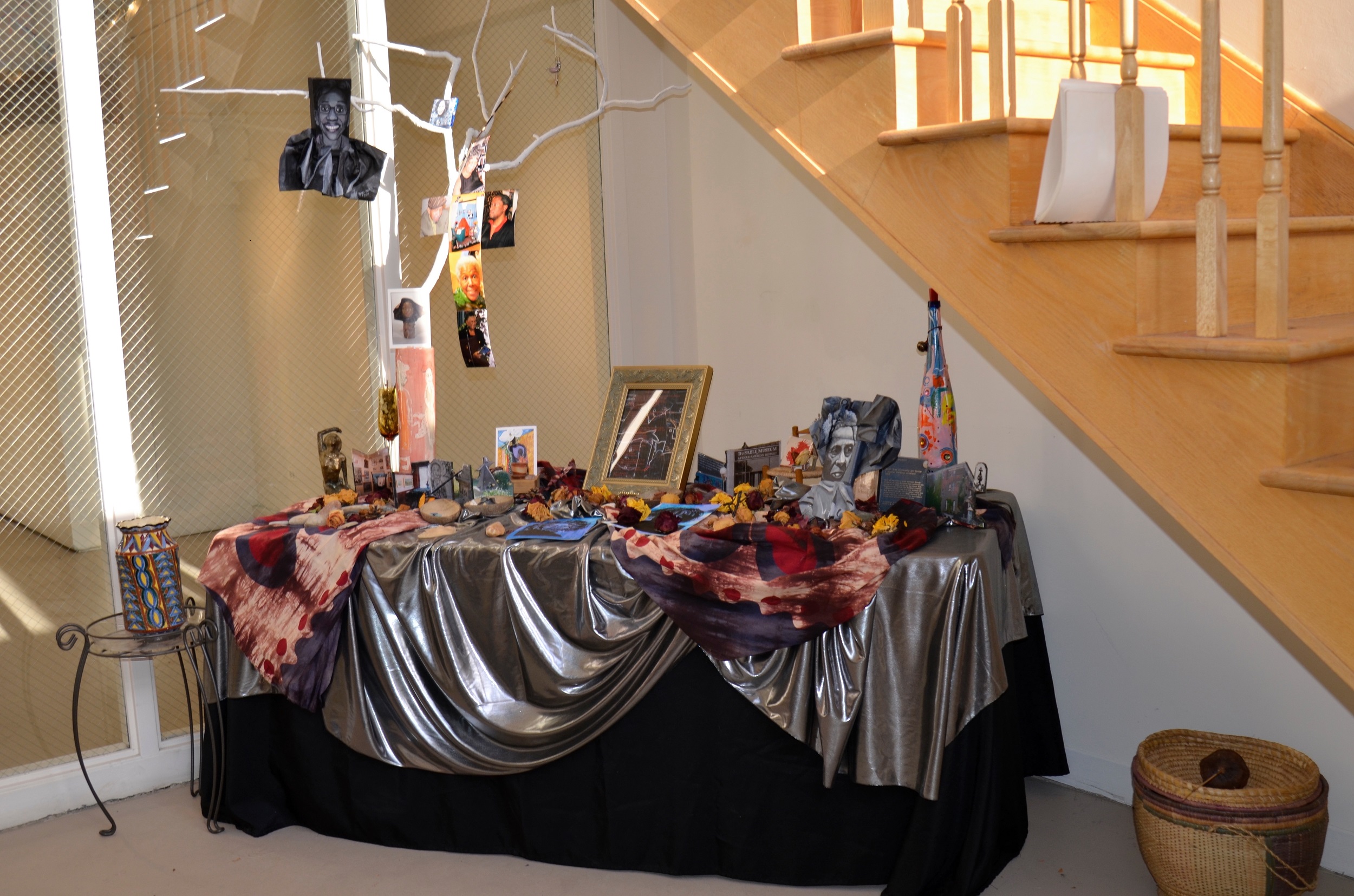 Color photograph of a table in front of a staircase, covered with a metallic cloth and various objects, including a tree branch hung with photographs