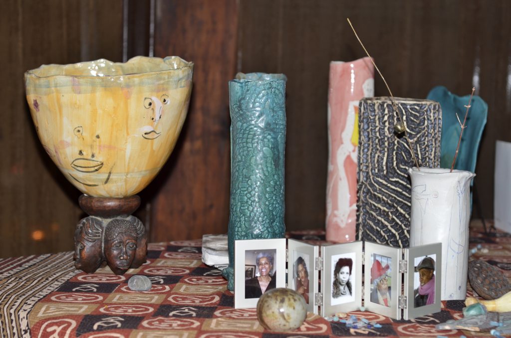 Color photograph of several candles, a yellow bowl with faces drawn on it, and a folding frame with five photographic portraits