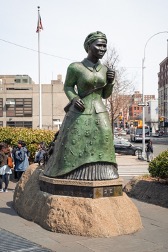Bronze statue sited in an urban setting of a woman in a long dress and headwrap striding forward