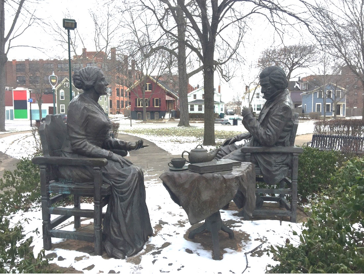 Bronze statue cited outdoors showing a woman and man seated by a small table with a teapot, cups, and books