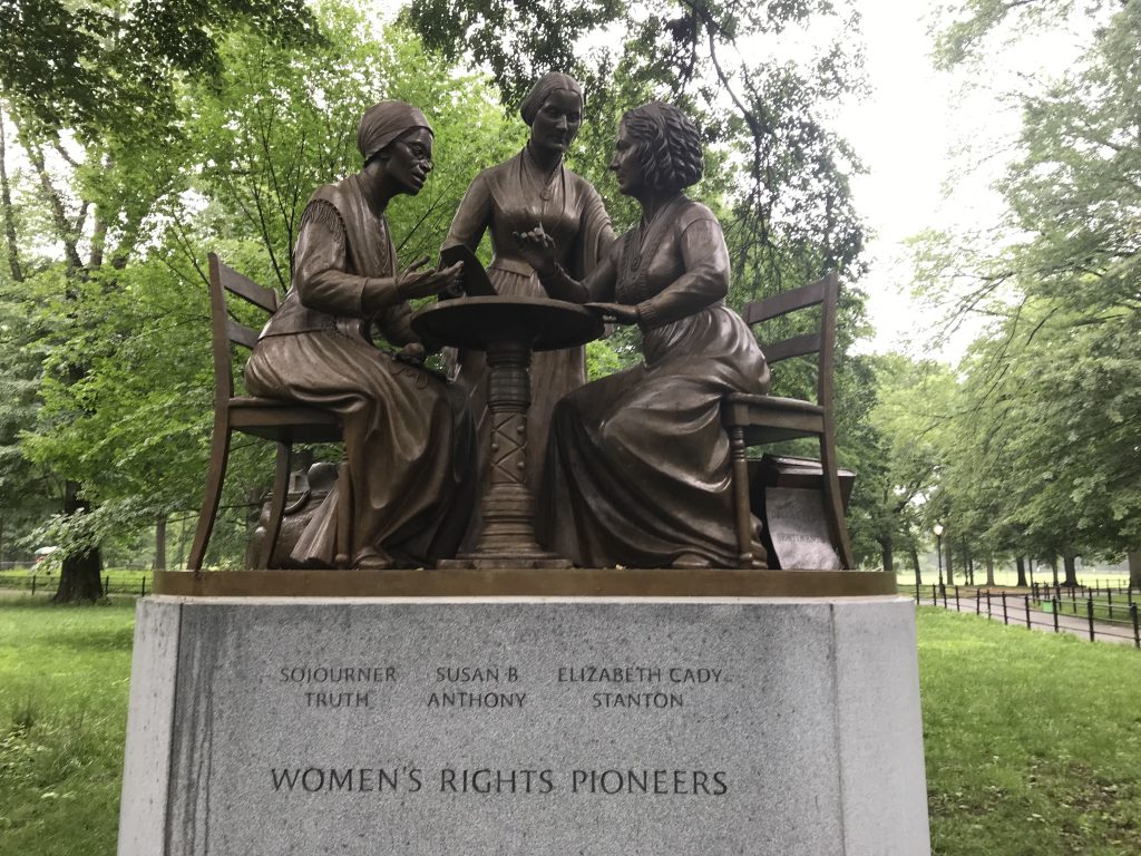 Bronze statue sited outdoors featuring three women in long skits seated at a table. The statue is on a stone plinth that reads WOMEN'S RIGHTS PIONEERS and provides the names of the women: Sojourner Truth, Susan B. Anthony, and Elizabeth Cady Stanton
