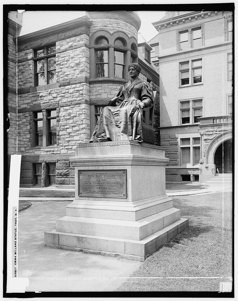Black and white photograph of a bronze statue of a seated woman on a stone pedestal, sited outside a stone building