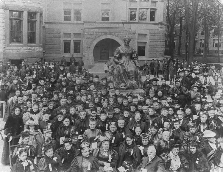 Black and white photograph of a large crowd gathered around a bronze statue of a seated woman, sited outside in front of a stone building