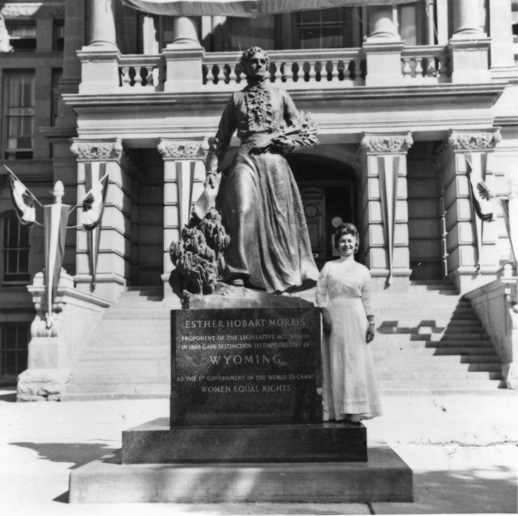 Black and white photograph of a bronze statue of a woman in a long skirt sited outdoors in front of a stone building. A woman in a long white dress stands next to the statue's pedestal, which reads ESTHER HOBART MORRIS