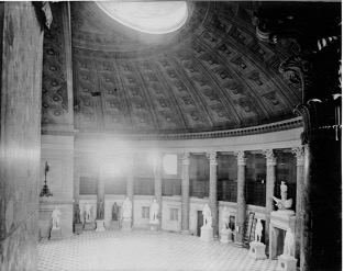 Black and white photograph of the interior of a rotunda with a coffered ceiling. Full-length statues of individuals surround the arcade.