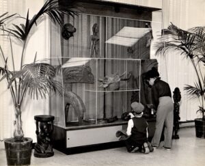 Black and white photograph of two young people looking into a glass display case filled with sculptural objects