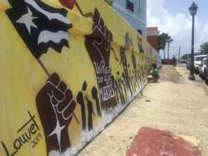 Photograph of a mural along a sidewalk showing hands holding a Puerto Rican flag against a yellow background. A figure in a green shirt crouches on the ground at the far end of the mural. 