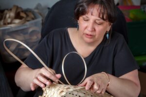 Color photograph of a dark-haired woman in a black top and turquoise earrings weaving a basket