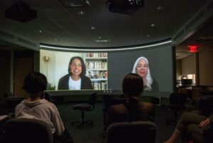 Projected screen of two women speaking side by side with an audience seated before them
