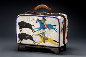 Color photograph of a suitcase with beaded decoration of two Native American horsemen pursuing two buffalo. The suitcase is inscribed "1903."