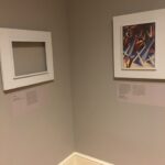 Interior view of a corner of a museum gallery with a work of art on the right side of the corner and an empty frame on the other side