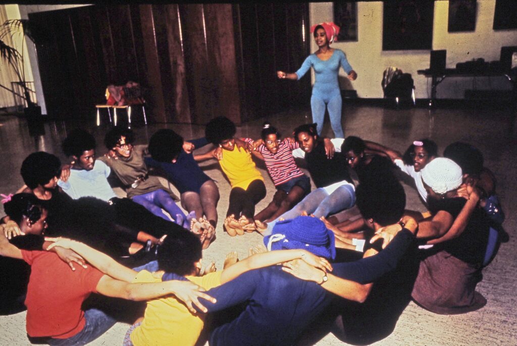 Color photograph of a group of people dressed in leotards, seated on the floor in a circle, their arms around each others' shoulders. One figure in a blue leotard and tights stands in the background.