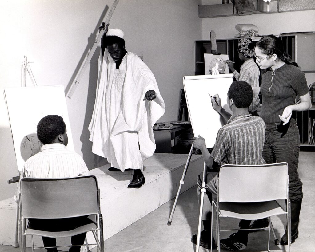 Black-and-white photograph of a life drawing class, with four people studying a live African American model in a white robe, holding a stick