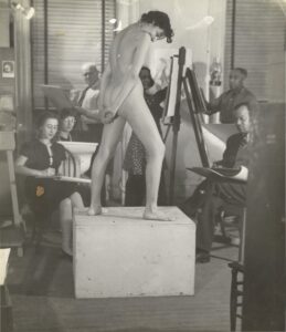 Black-and-white photograph of a nude female model being sketched by six students