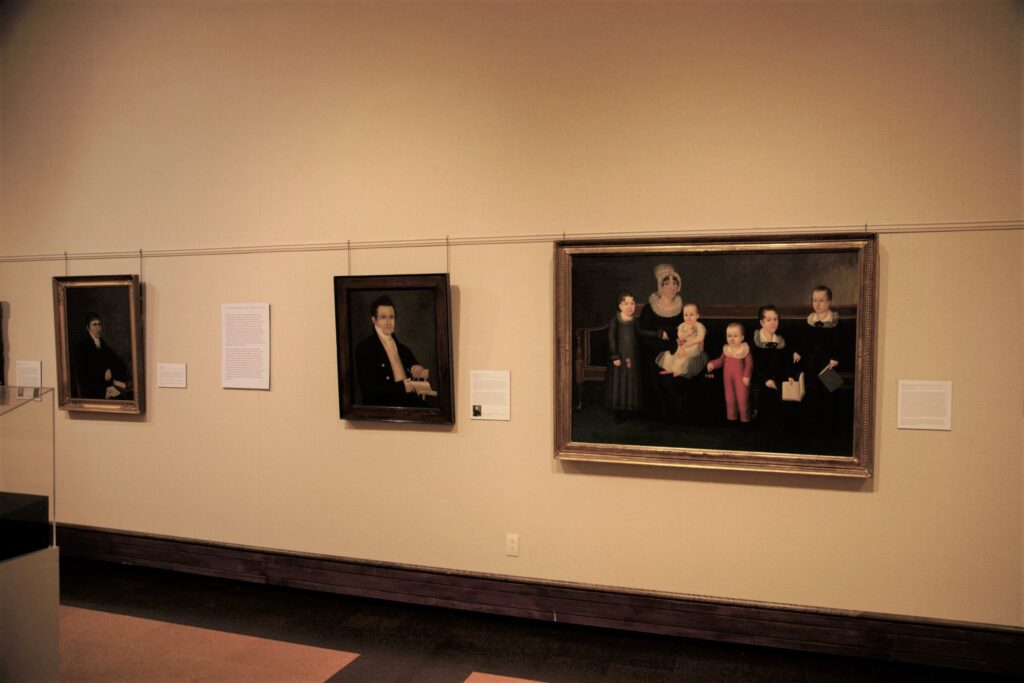 Interior view of a museum gallery wall with three paintings; the one on the far right is of a woman and five children