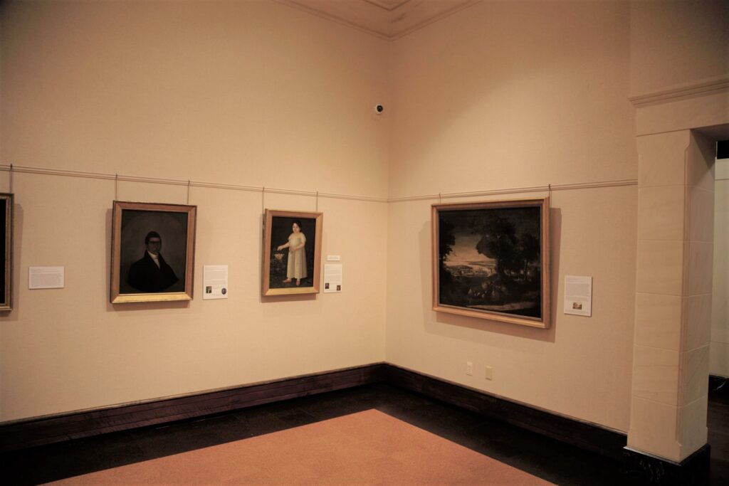 Interior view of a corner of a museum gallery with three paintings and a partial doorway visible on the right