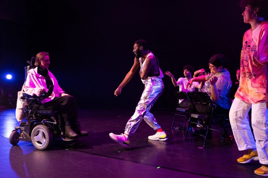 On a dark stage with purple lighting, Jerron Herman, a dark-skinned Black man wearing a silver jumpsuit, dances with Park McArthur, a white woman in a power wheelchair, as Carolyn Lazard, a light-skinned Black nonbinary person, stands behind. Seated audience bops along.