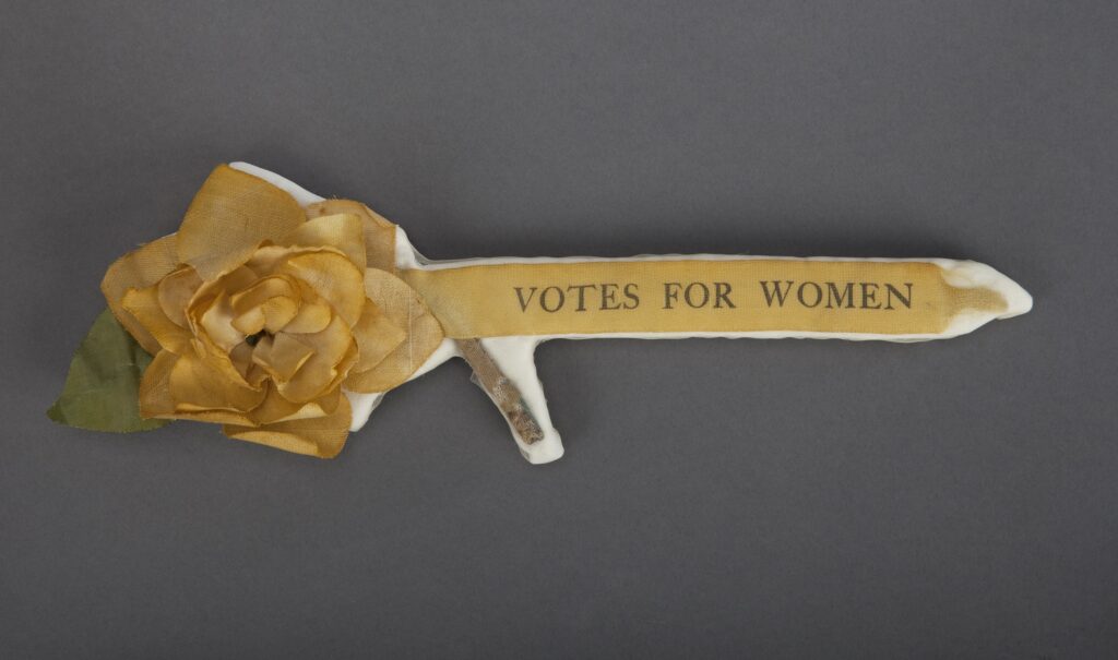 Photograph of a yellow stickpin with a sunflower shape on one end, reading "Votes for Women"