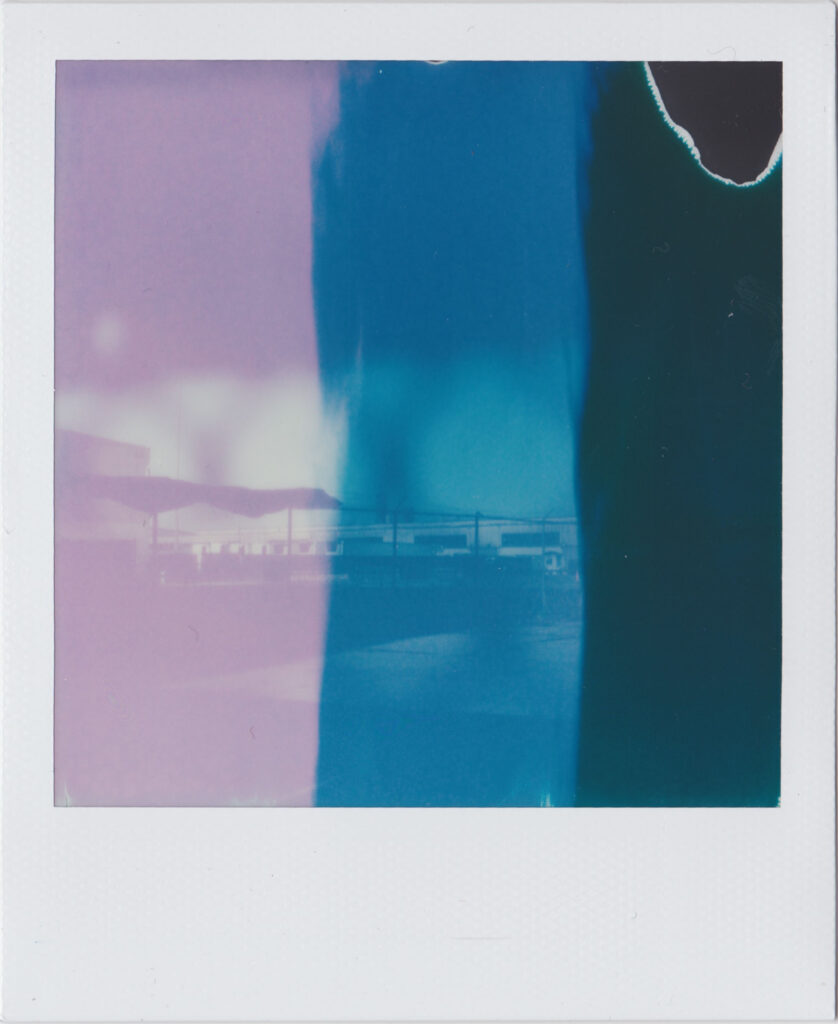 Polaroid photograph of distant low buildings; an exposure effect has superimposed vertical stripes of mauve, blue, and black over the image.