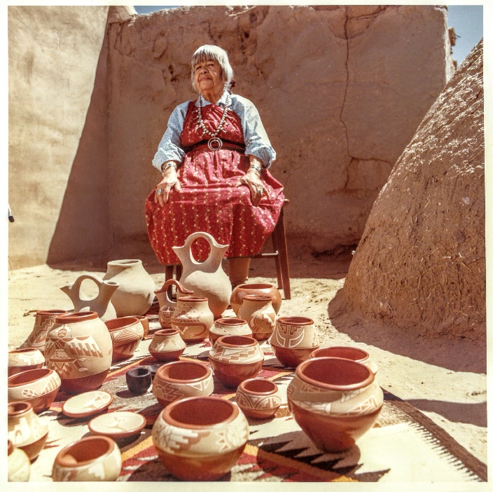 Color photograph of an elderly woman in a red dress sitting in front of an adobe structure, with dozens of handmade pots in front of her