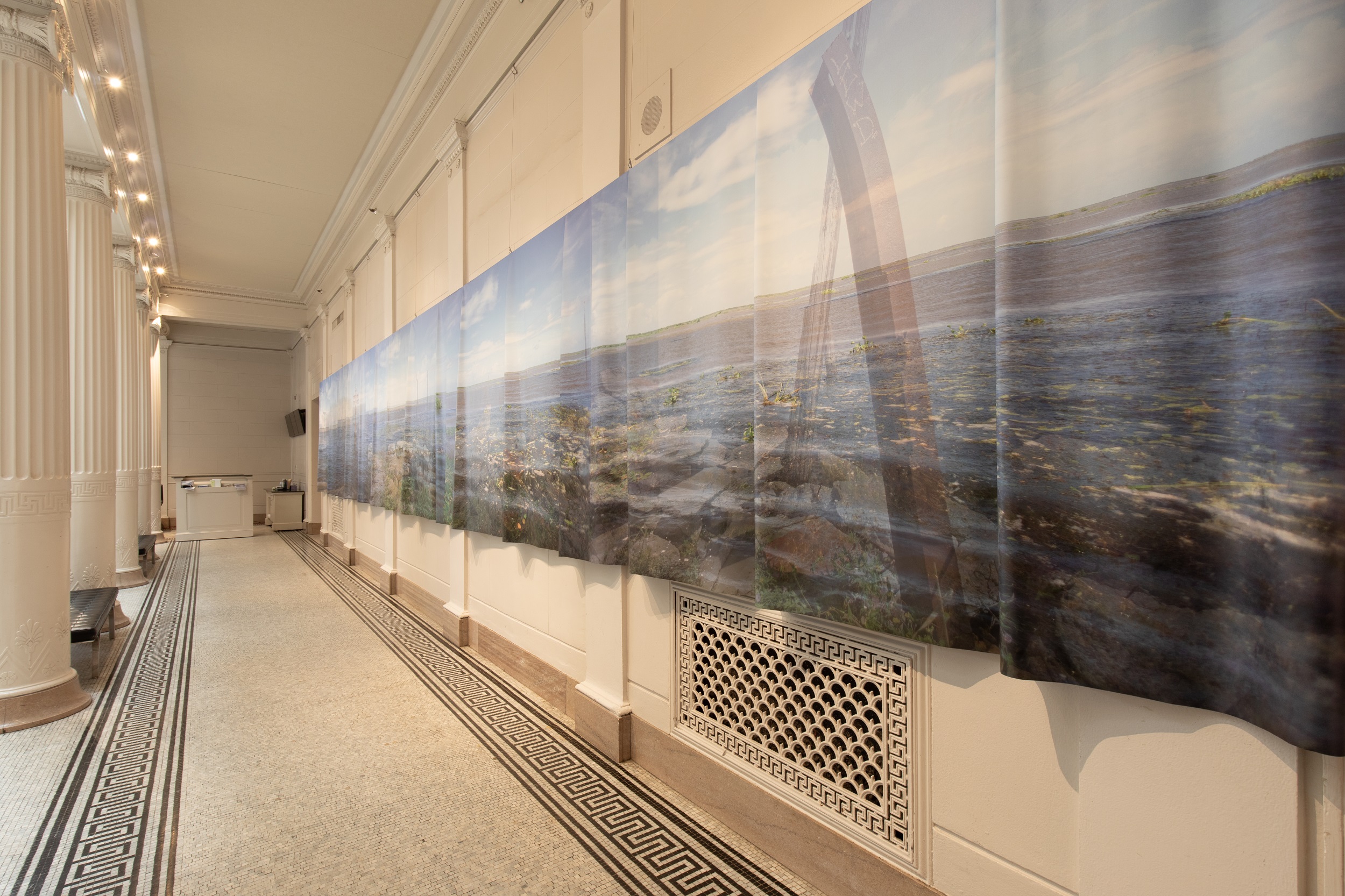 A fabric painting of an industrial waterscape hung in the interior hallway of a white, neoclassical building