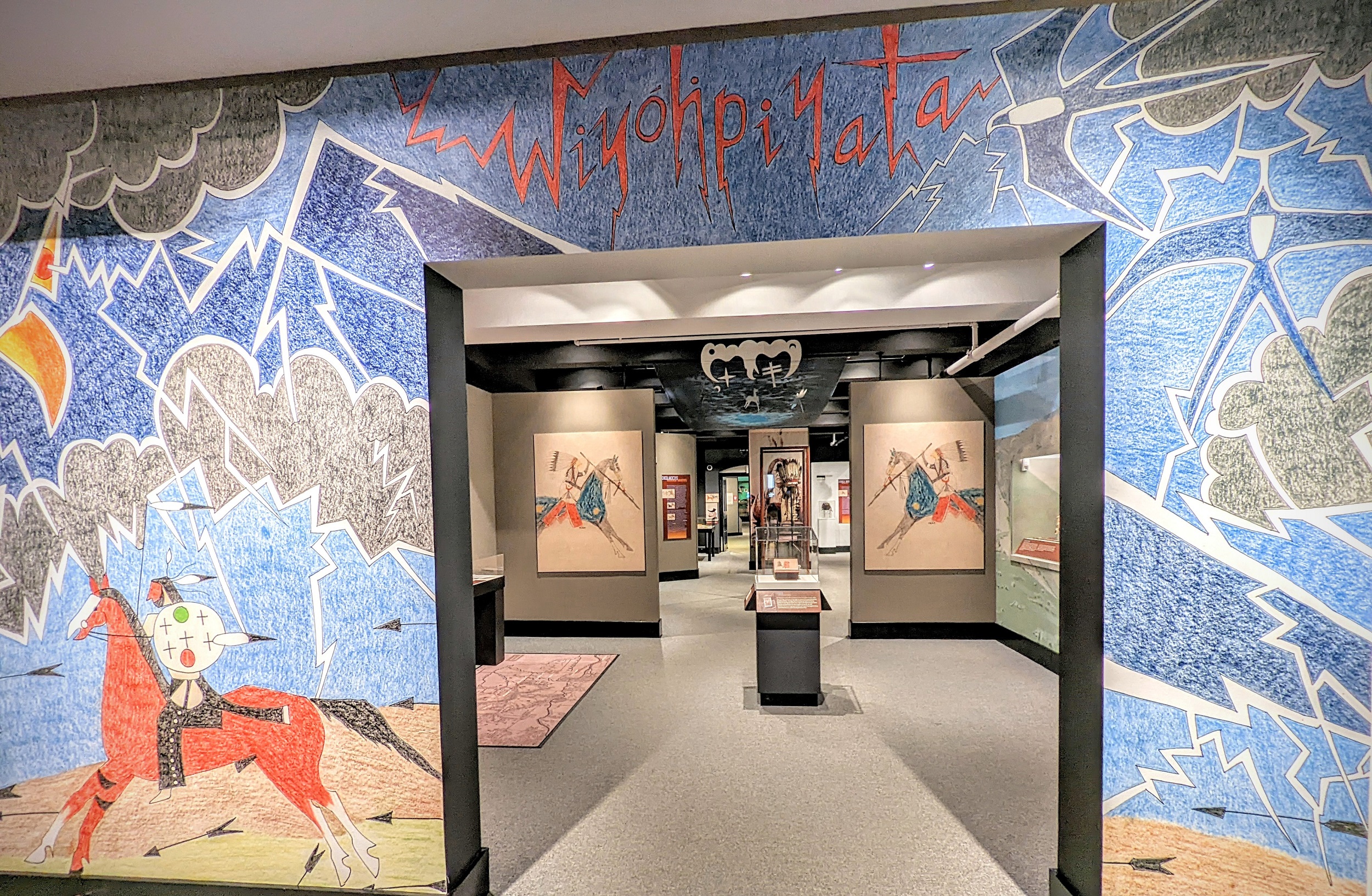 Entrance to a museum gallery, with the entrance wall decorated in jagged landscape forms, with a horse and rider on the left, birds on the right, and the word "Wiyohpiyata" above the doorway