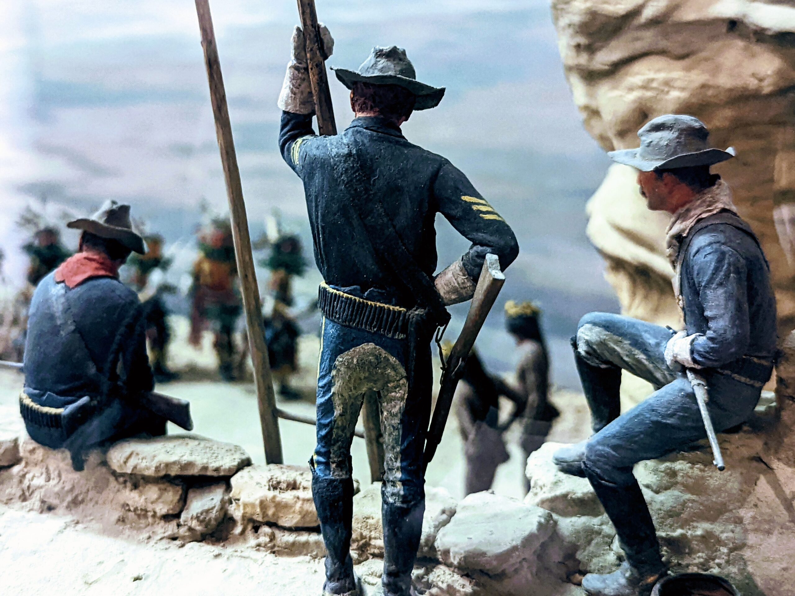 Three men in US Cavalry uniforms, seated and standing in a rocky landscape, watch a group of Native Americans just beyond
