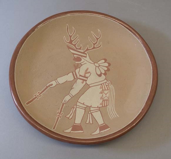 Ceramic plate with central figure in Native American dress, wearing deer antler on their head and holding a stick in each hand
