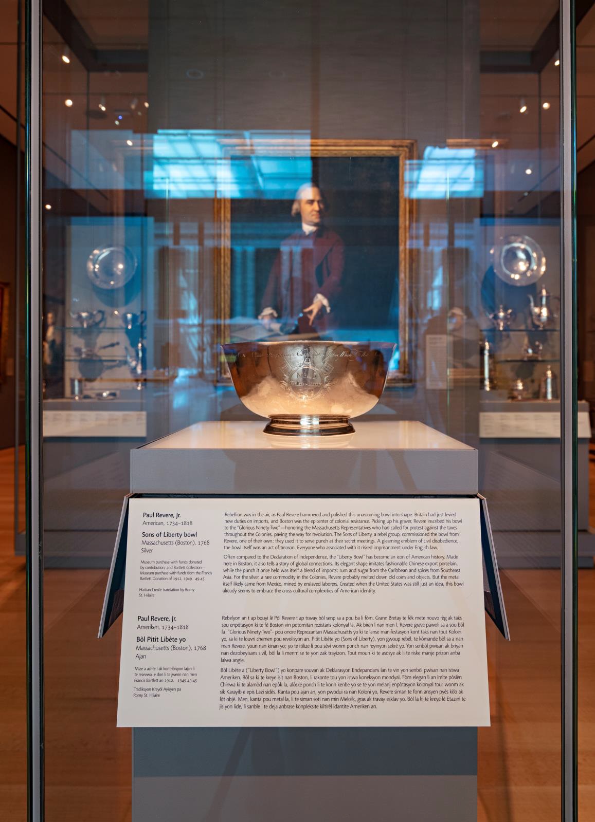 Close up on a silver bowl in a glass case in a museum gallery, with a portrait of a man in colonial American garb seen through the case
