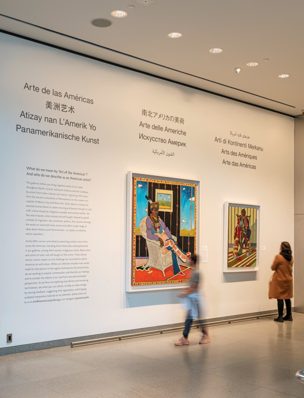 Photograph of entrance wall to museum exhibit gallery, with two figures standing in front of two paintings and a text panel, over which is the title "Arte de las Americas" in several languages