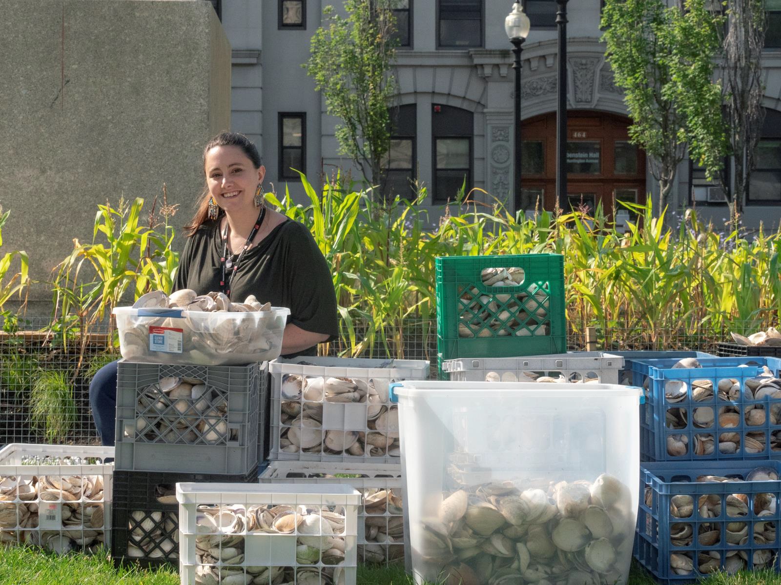 Color photograph of a smiling woman seated behind bins of seashells in an urban garden setting