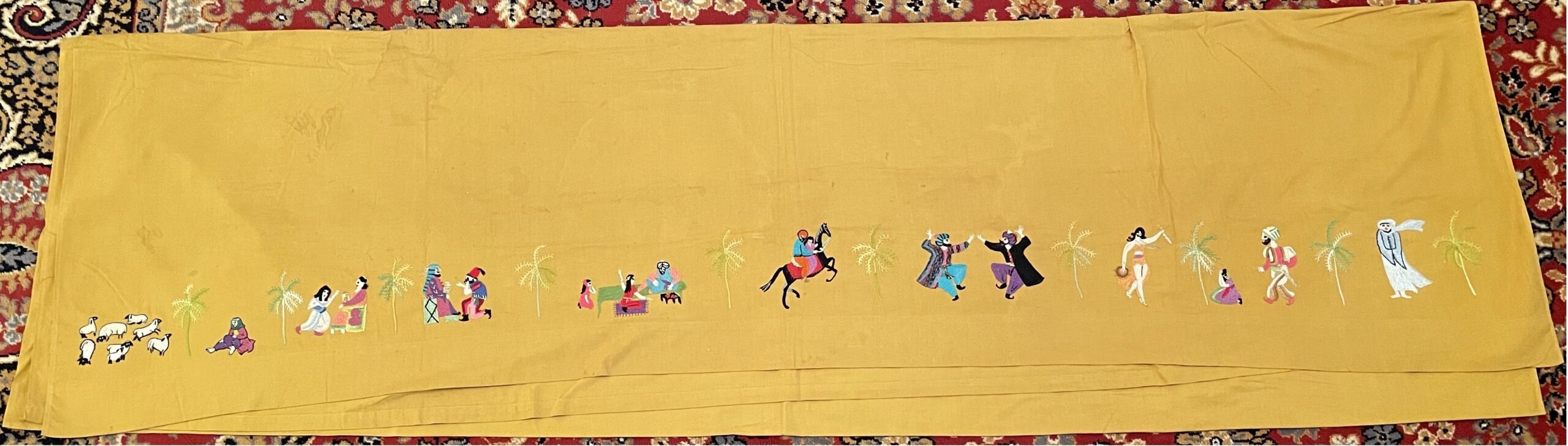 Color photograph of a horizontal piece of cloth, yellow-orange in color, embroidered with a line of tiny people, animals, and trees along the bottom edge