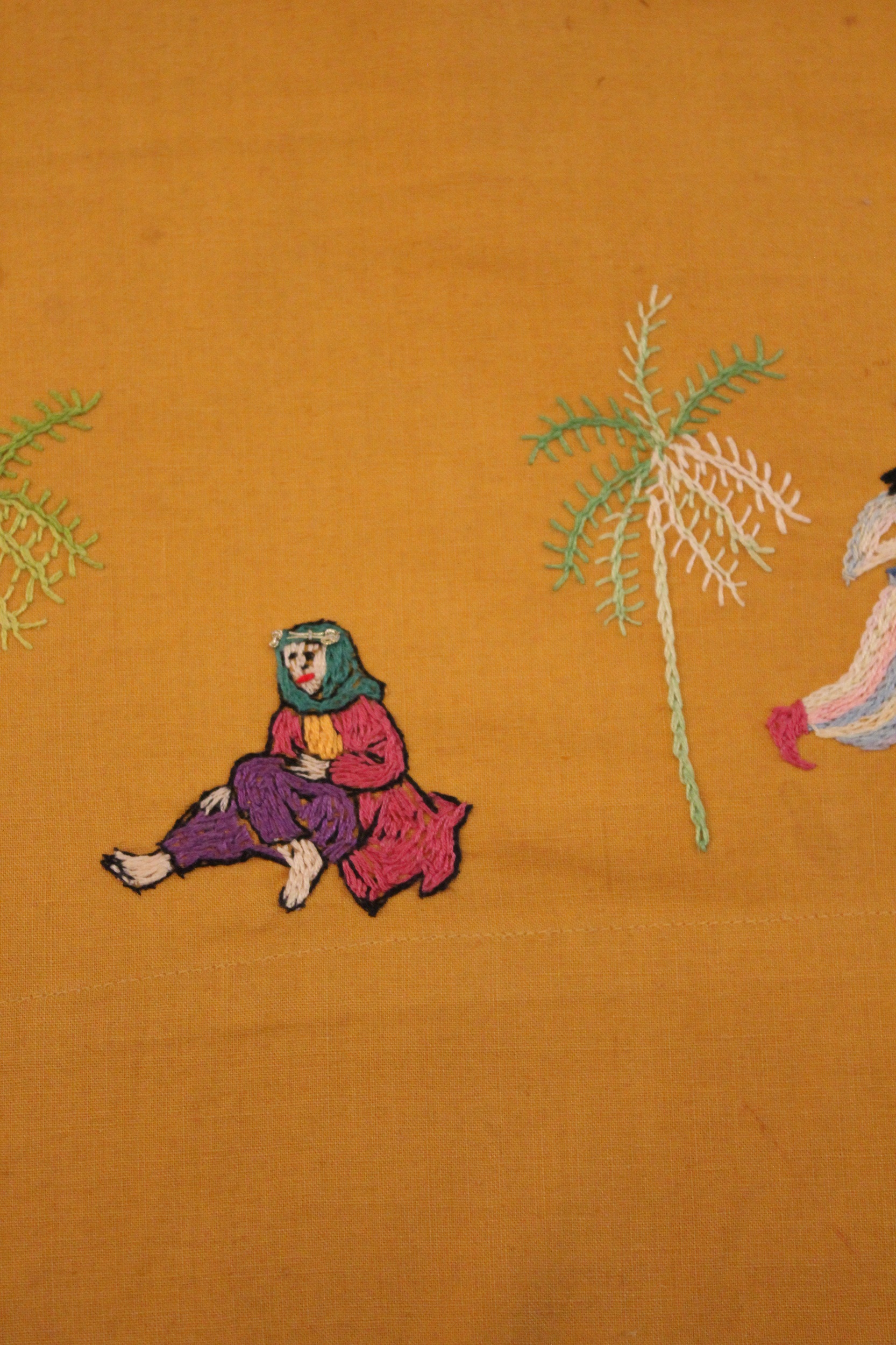 Embroidered depiction of a figure in purple pants, a red coat, and a green headscarf sitting on the ground, between two palm trees