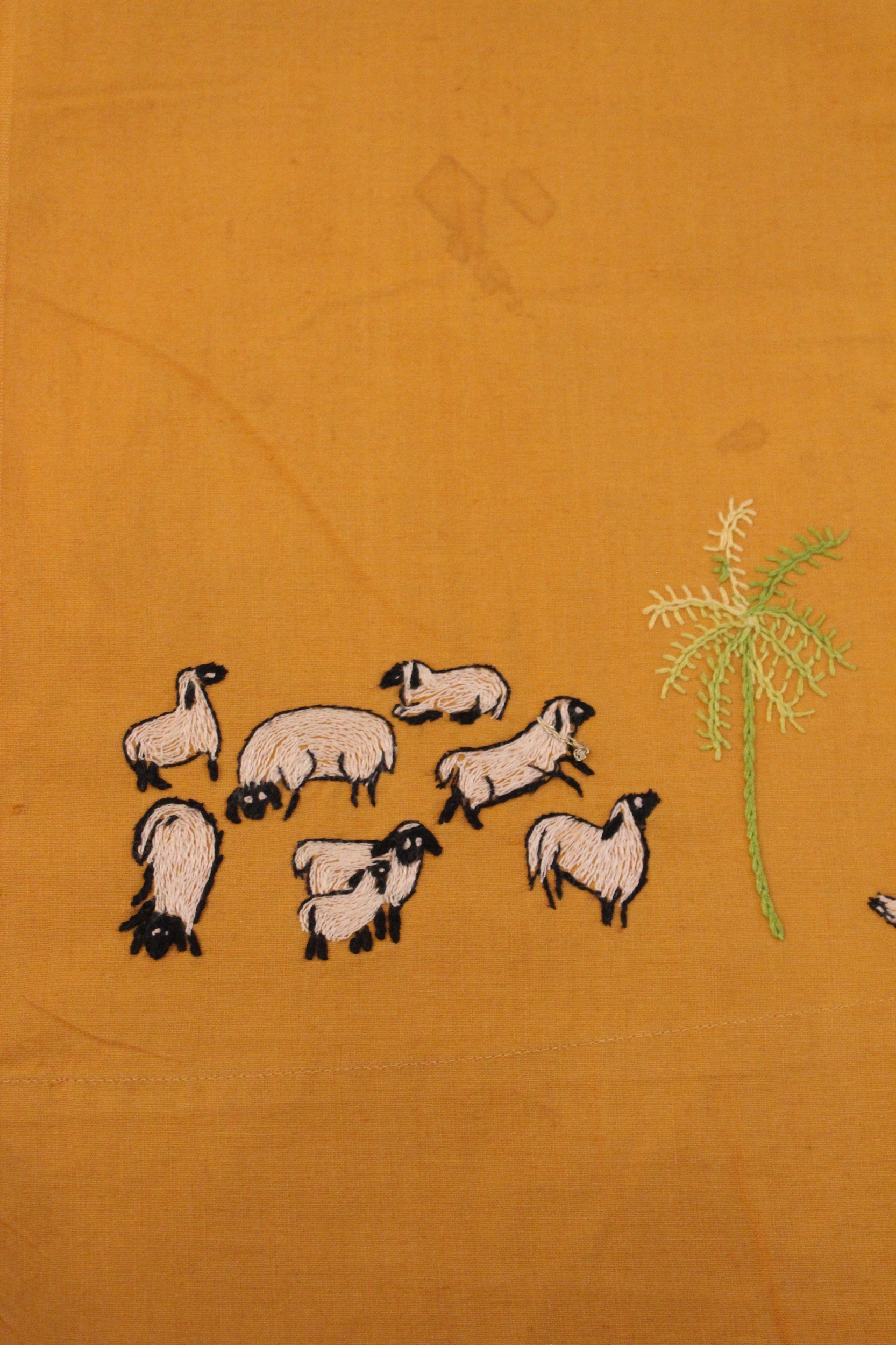 Embroidered depiction of eight sheep with white bodies and black faces, ears, and feet. A small palm tree is to the right.