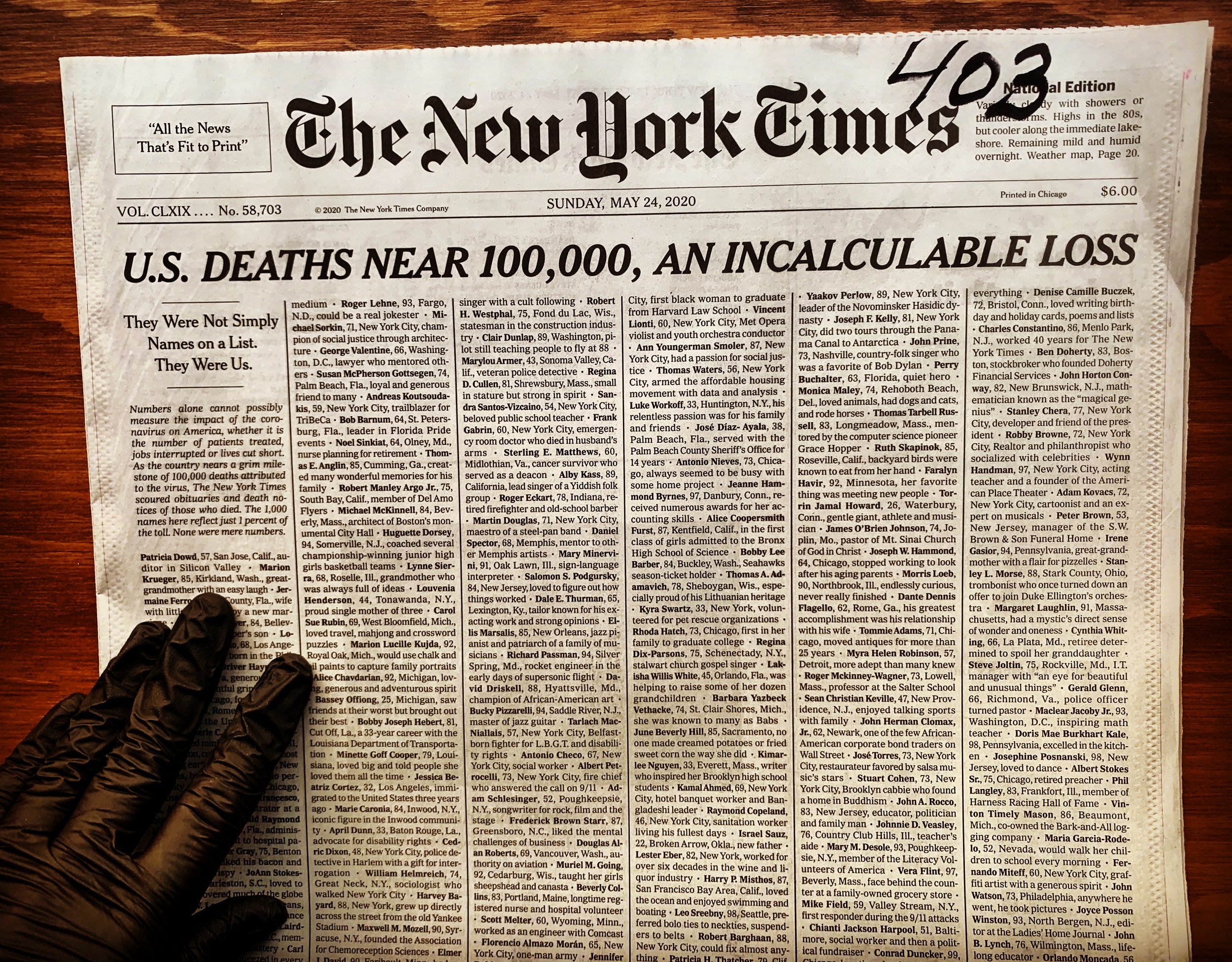 Color photograph of the front page of the New York Times with the headline "U.S. Deaths Near 100,000, an Incalculable Loss." A gloved hand appears in the lower left corner of the image, near the fold of the newspaper.