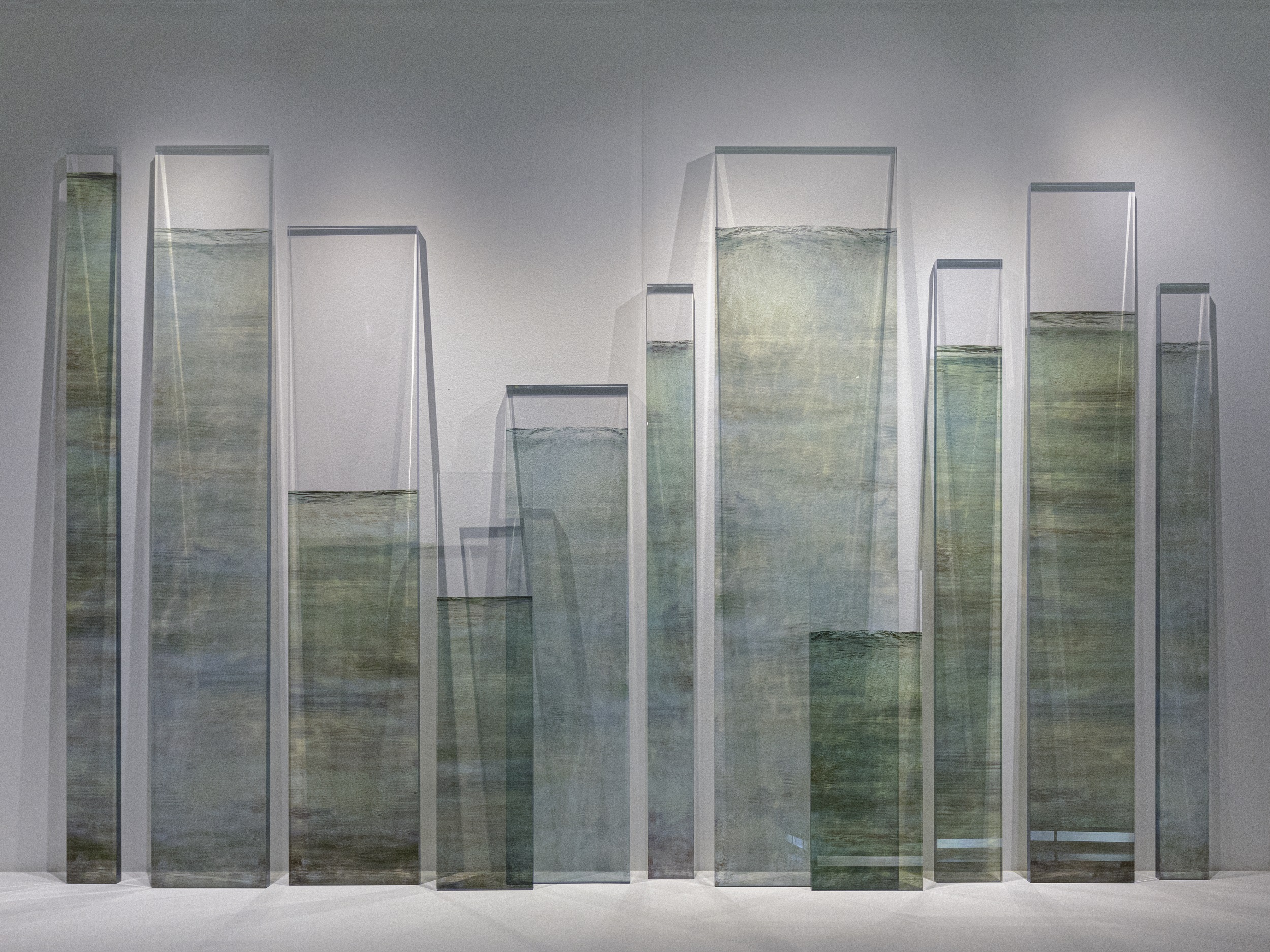 Photograph of artwork, composed of several vertical, translucent rectangles in shades of gray, green, and blue, resting against a white gallery wall