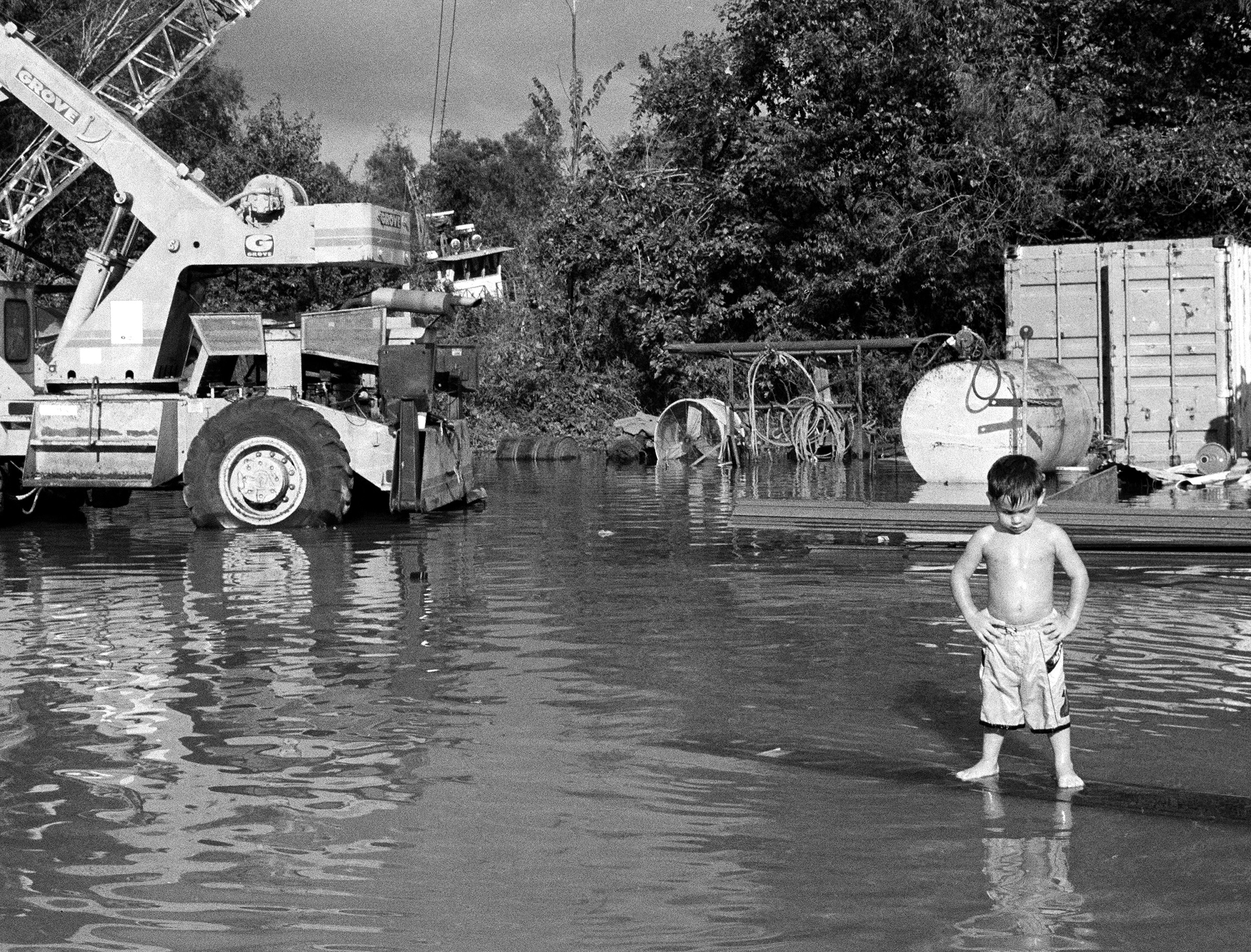 Black-and-white photograph of a young boy in shorts standing in a large puddle, posing in front of a construction vehicle