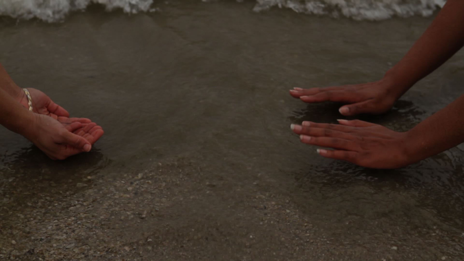Photographic image of two sets of hands, one lifting up and the other pushing down, at the sand near the edge of the waves