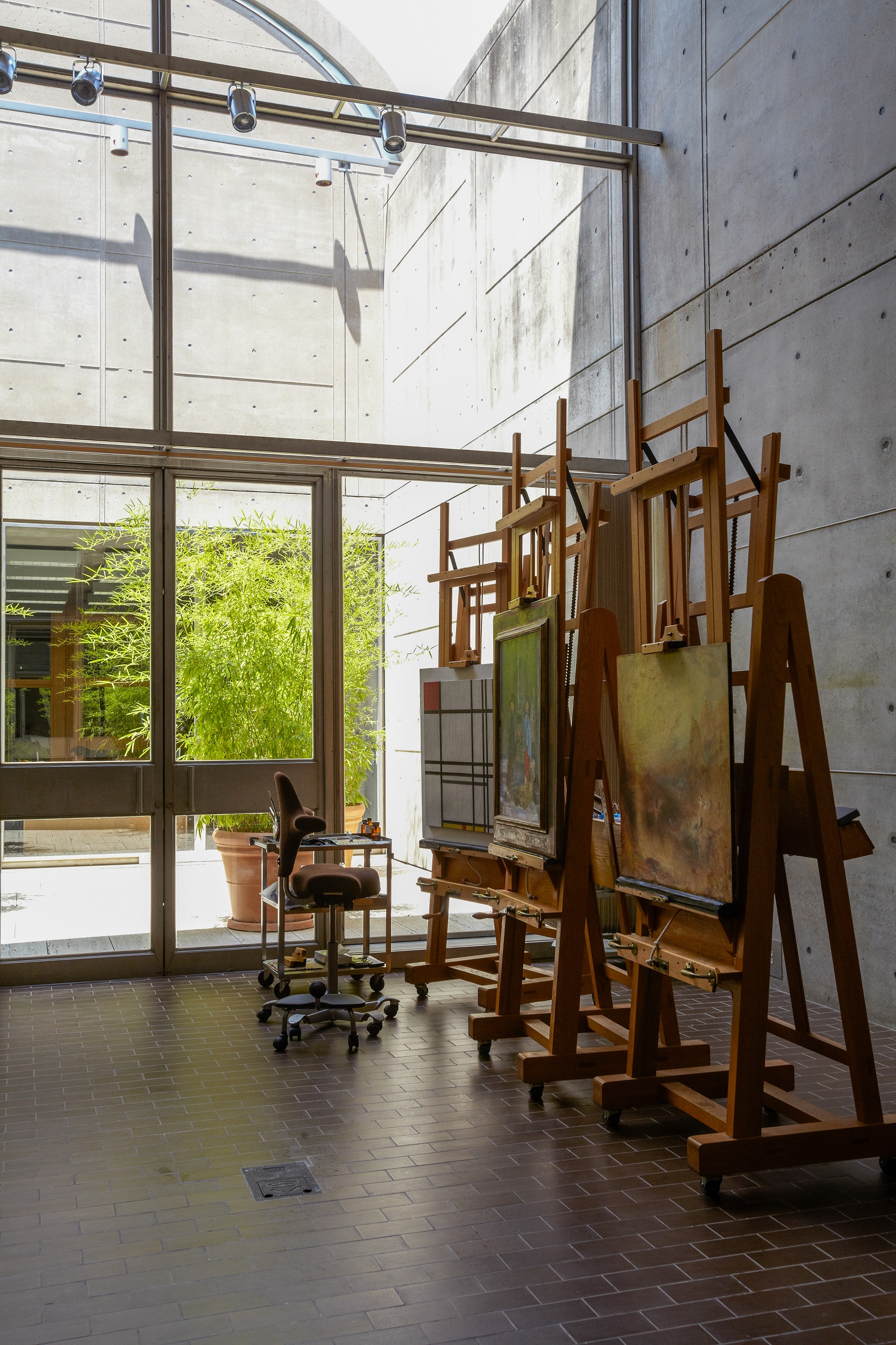 Interior of a naturally lit room with a large glass wall on the left and easels with paintings on the right.