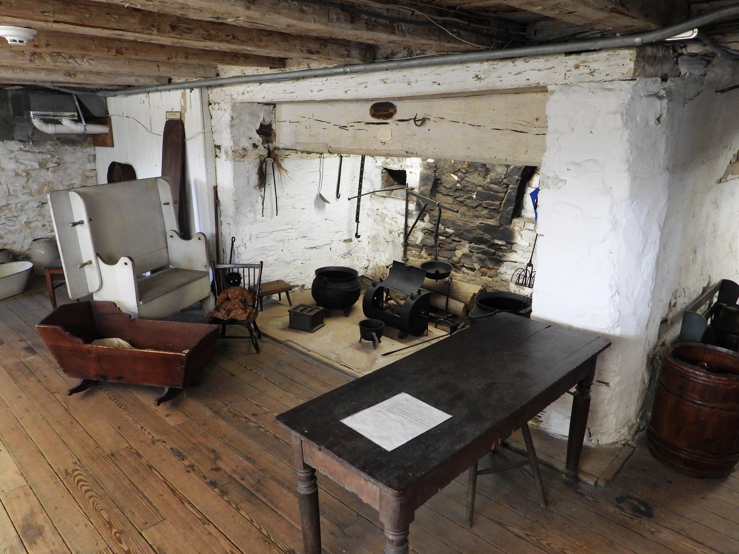 Interior of a replica colonial American basement kitchen, with a wide, whitewashed fireplace, a high-backed bench, a wooden cradle, and a table in front