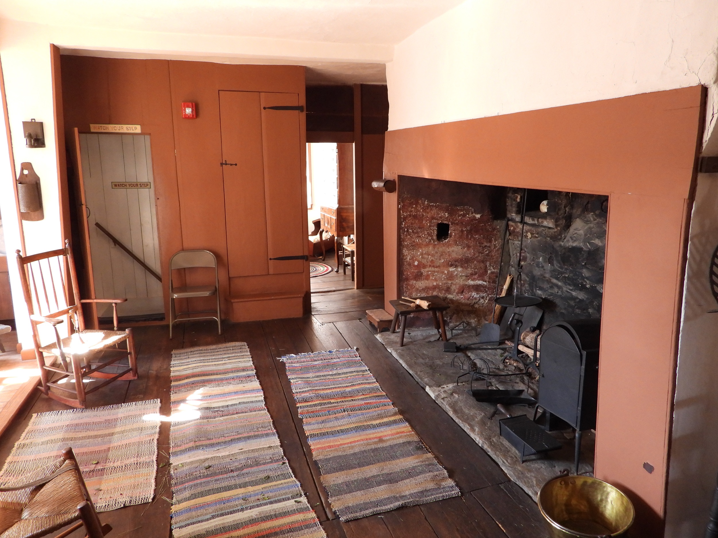 Photograph of a replica colonial American first-floor kitchen, with a wide fireplace with two rocking chairs and three woven rugs in front of it